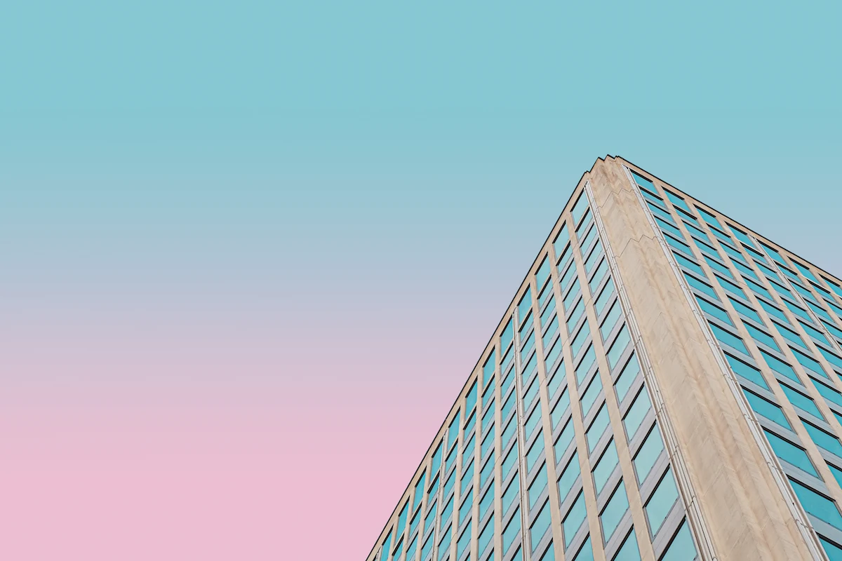 The upper corner of a building somewhere in London, UK. The building's facade is tan, possibly concrete or stone. Each wall has a grid of square shaped, bluish-green windows. Since this photo appears to have been shot at sunset, the sky ranges from blue at its top to a rosy orange below.