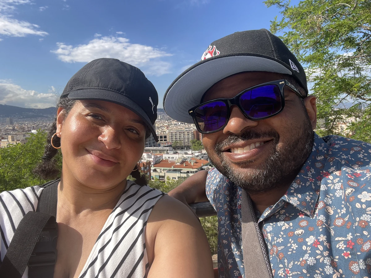 Jason and I in Barcelona. I am on the left, facing the camera wearing a black baseball-style cap and a sleeveless white dress with diagonal black stripes. Jason is also wearing a black baseball cap, with dark-framed glasses. He is wearing a floral-print shirt with a teal background and red and white flowers in the foreground.