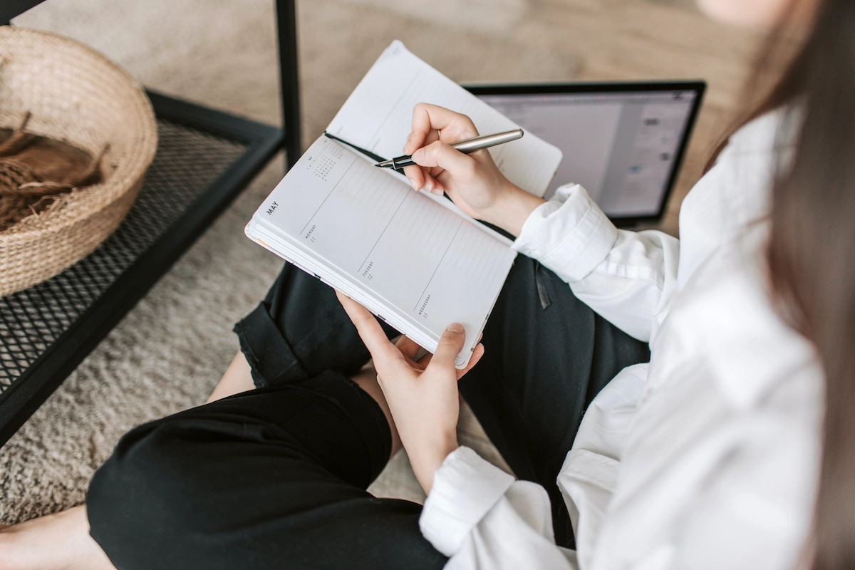 A feminine-presenting person sits wearing a white shirt and black pants sits cross-legged on a cream-colored carpet. They're holding a weekly planner, opened to the week of May 11. They're about to write something in the planner using the fountain pen they're holding in their right hand.