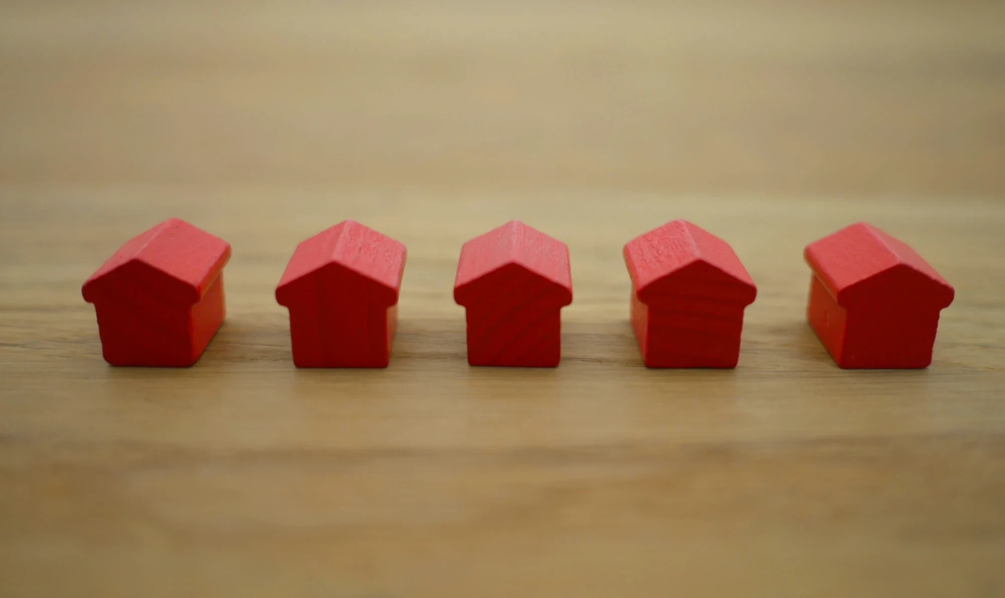 Four small red wooden blocks shaped like houses rest on a oak-colored wooden table surface.